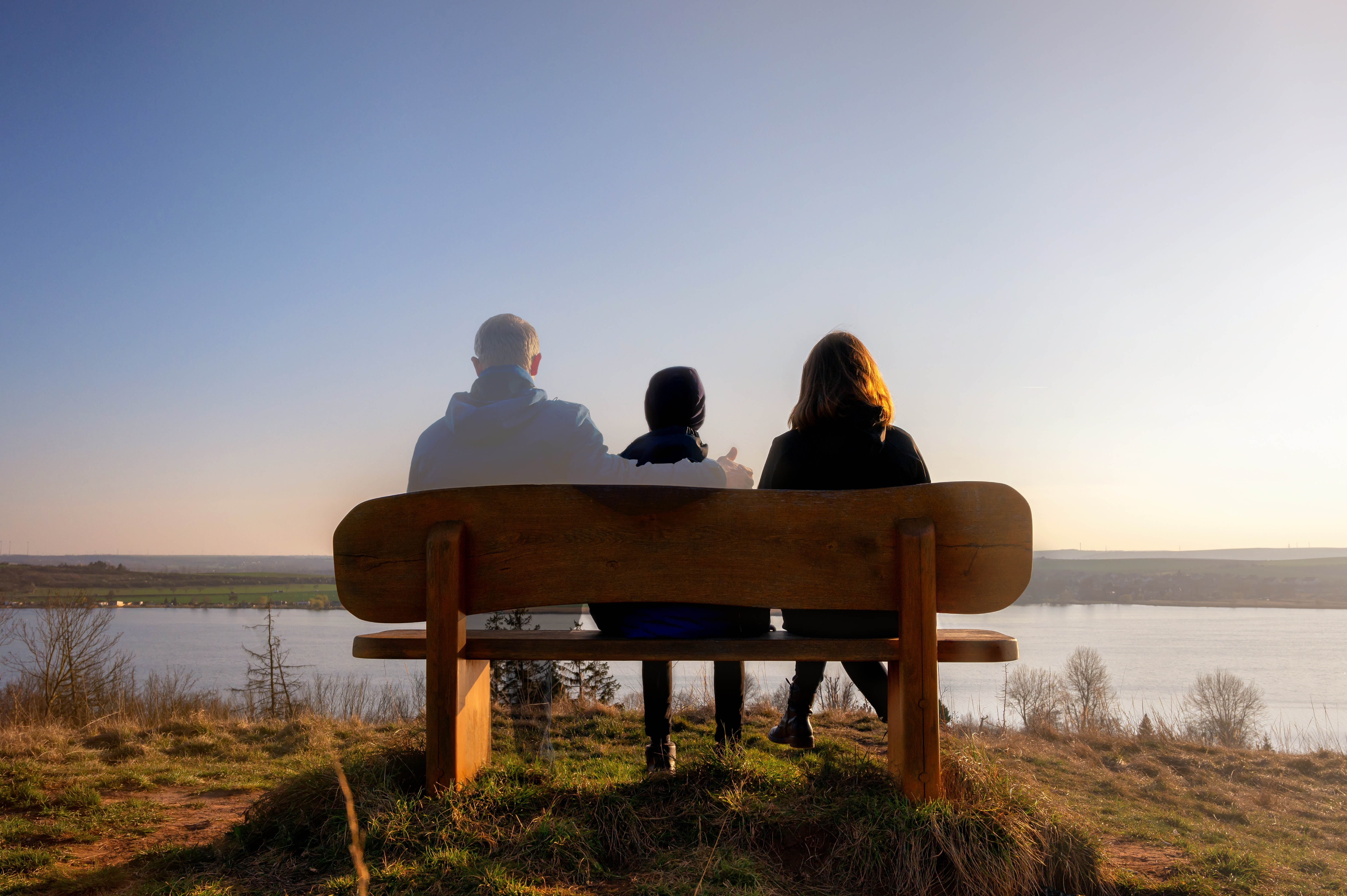 Child and mom sitting on a bench looking out to the water, with a faded silhouette of the dad - showing mourning the loss of a parent Child and mom sitting on a bench looking out to the water, with a faded silhouette of the dad - showing mourning the loss of a parent